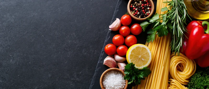 Italian cooking ingredients arranged along the right edge of a dark slate surface with copyspace on the left
