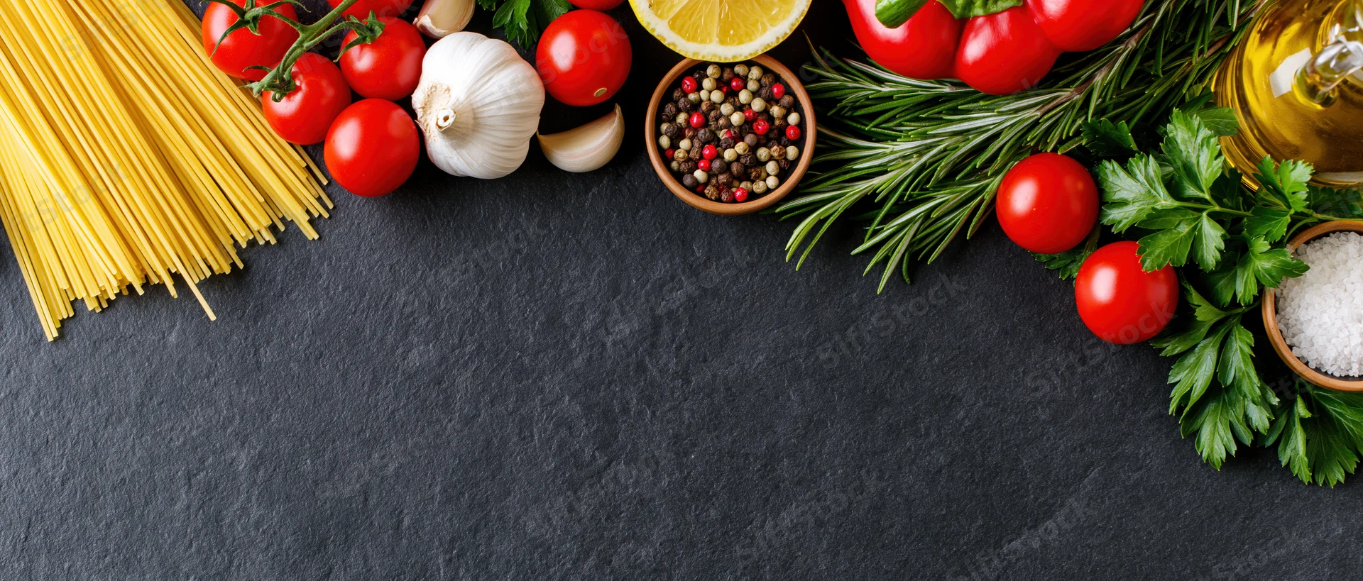 Italian cooking ingredients arranged along the top edge of a dark slate surface with copyspace below