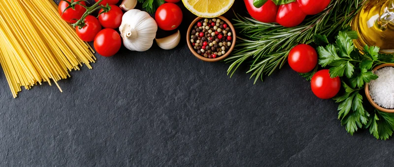 Italian cooking ingredients arranged along the top edge of a dark slate surface with copyspace below