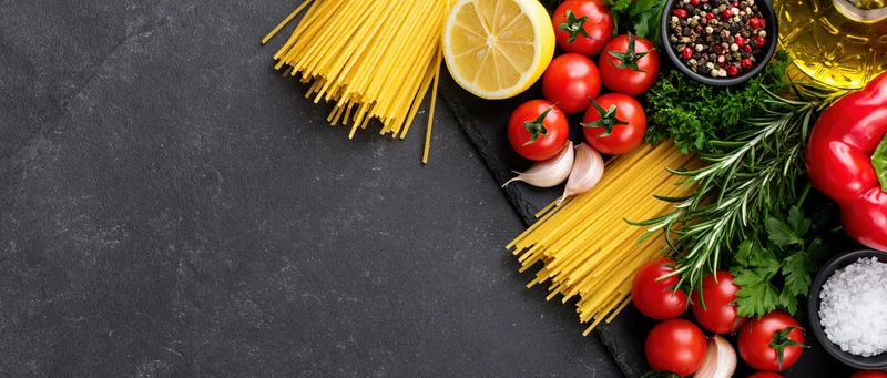 Italian cooking ingredients arranged along the right edge of a dark slate surface with copyspace on the left
