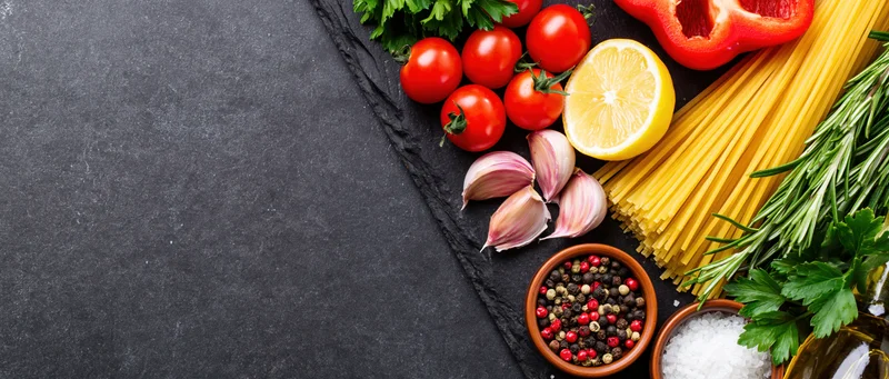 Italian cooking ingredients arranged along the right edge of a dark slate surface with copyspace on the left