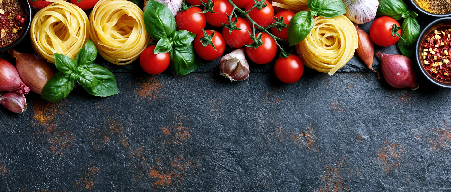 Italian cooking ingredients arranged along the top edge of a dark stone surface with copyspace below
