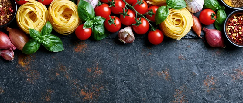 Italian cooking ingredients arranged along the top edge of a dark stone surface with copyspace below