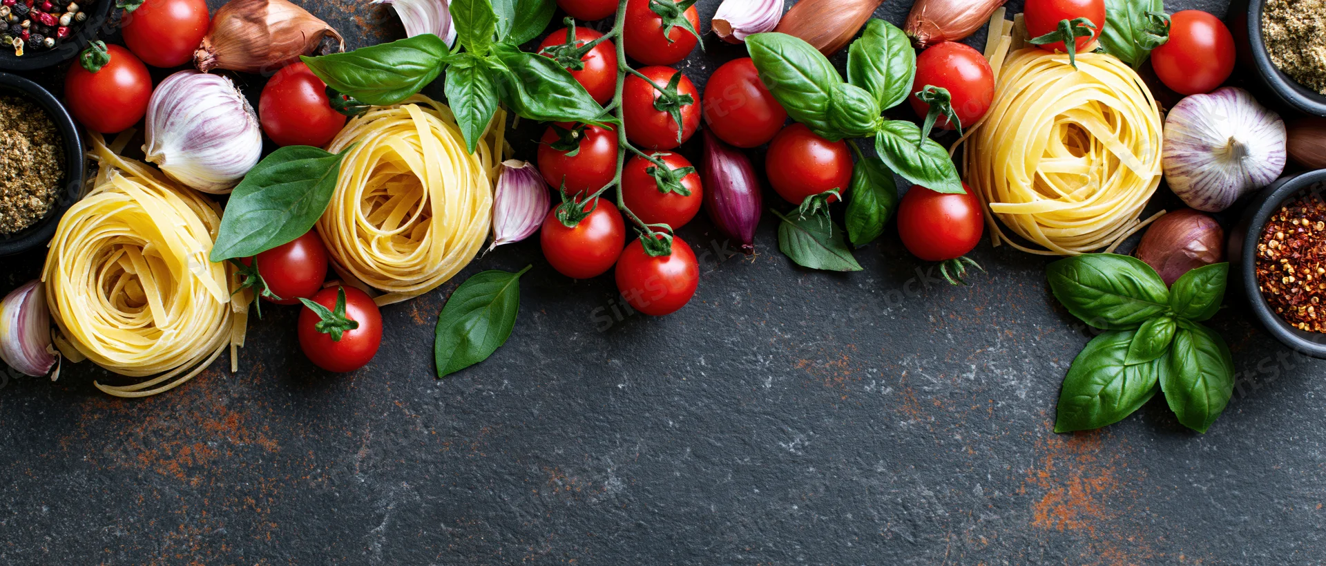 Italian cooking ingredients arranged along the top edge of a dark slate surface with copyspace below