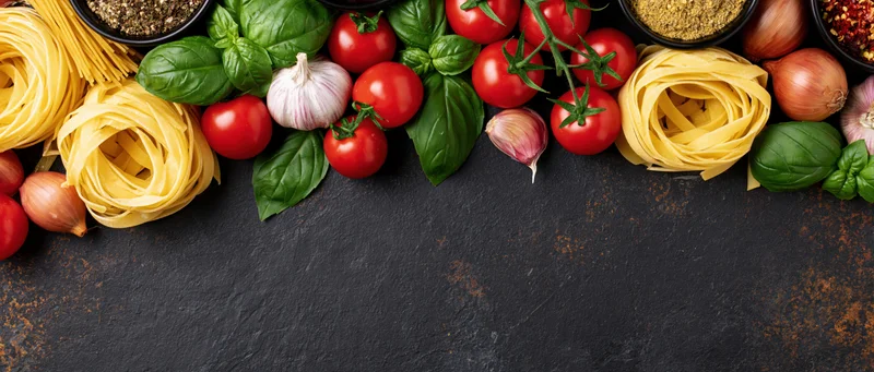 Italian cooking ingredients arranged along the top edge of a dark slate surface with copyspace below