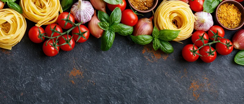 Italian cooking ingredients arranged along the top edge of a dark slate surface with copyspace below