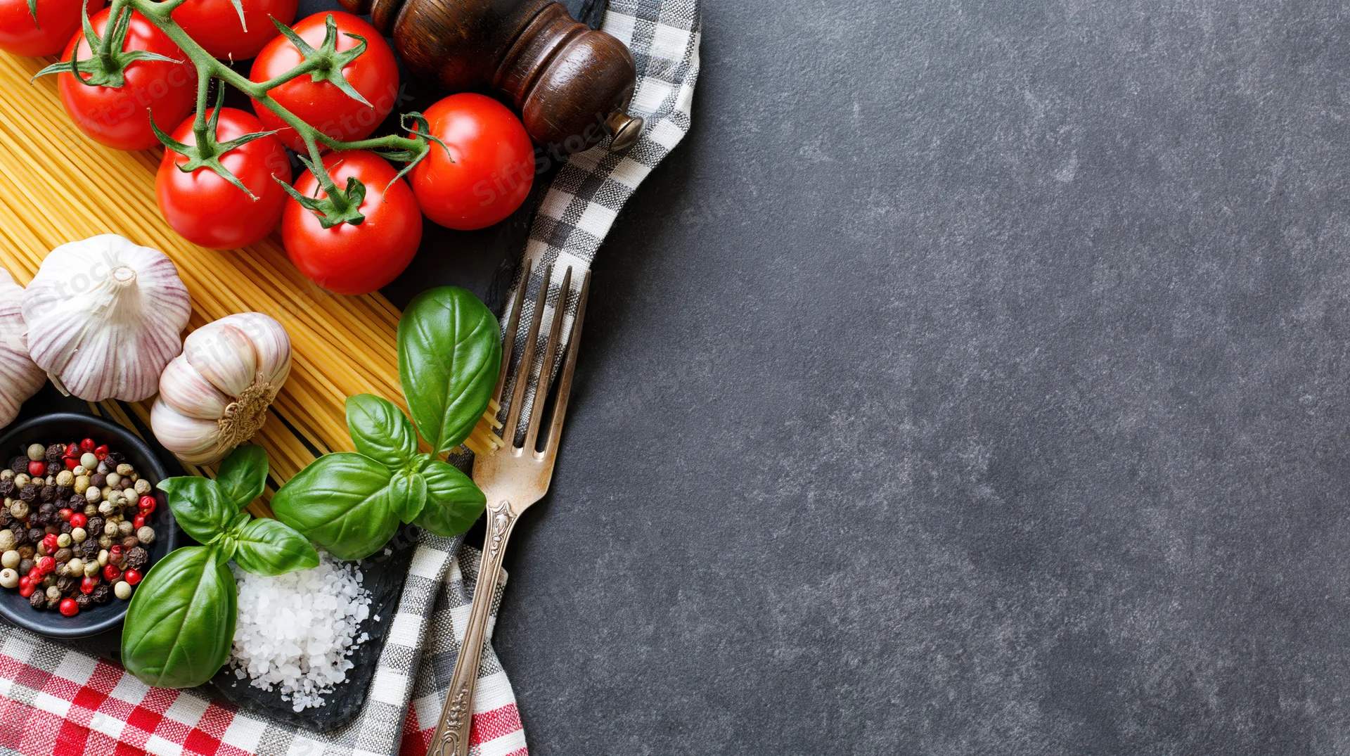 Italian cooking ingredients arranged on left side of dark stone countertop with checkered cloth and fork