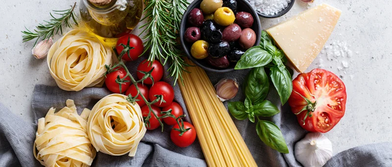 Italian cooking ingredients including pasta, tomatoes, basil, garlic, olive oil, olives and cheese arranged on a light countertop with a gray cloth
