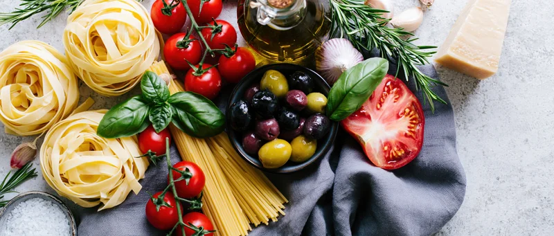 Italian cooking ingredients including pasta, tomatoes, basil, olive oil, garlic, cheese, and olives arranged on a light concrete surface with a gray cloth and copyspace on the right