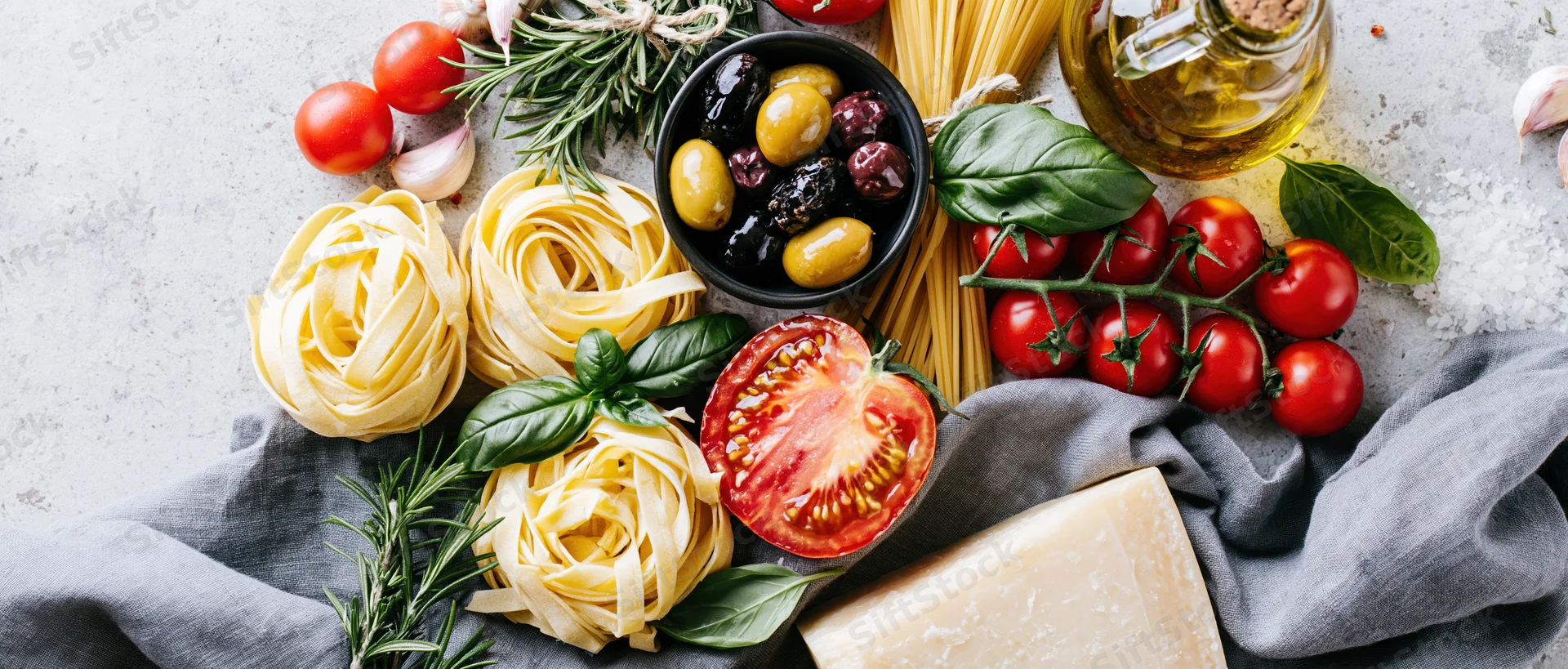 Italian cooking ingredients including pasta, tomatoes, basil, olives, garlic, rosemary, cheese and olive oil arranged on a light gray stone countertop with a gray cloth