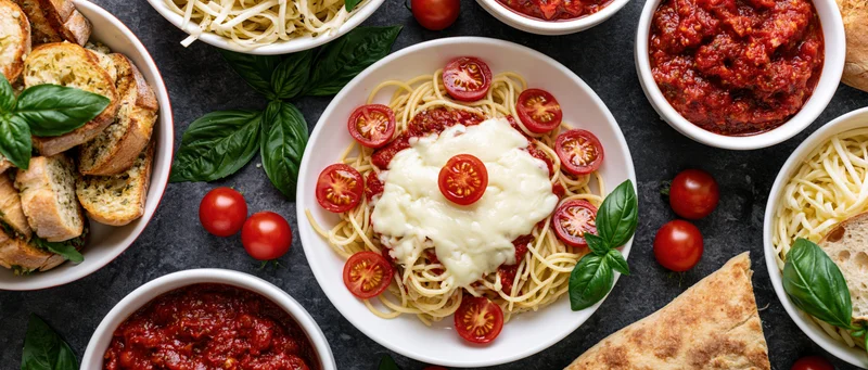 Spaghetti with tomato sauce and mozzarella topped with cherry tomatoes and basil on a dark slate table surrounded by garlic bread and side dishes