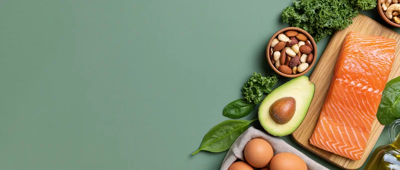 Keto diet food flat lay with salmon avocado eggs nuts kale and olive oil on wooden board against green background