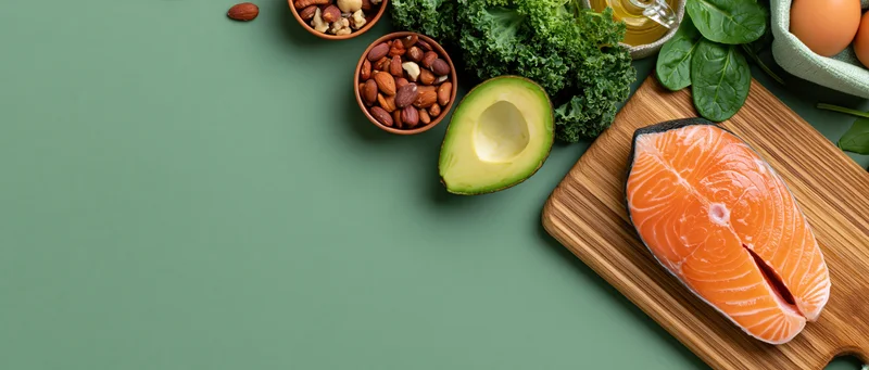 Salmon fillet and avocado on cutting board with nuts, kale, spinach, eggs and olive oil arranged on green surface