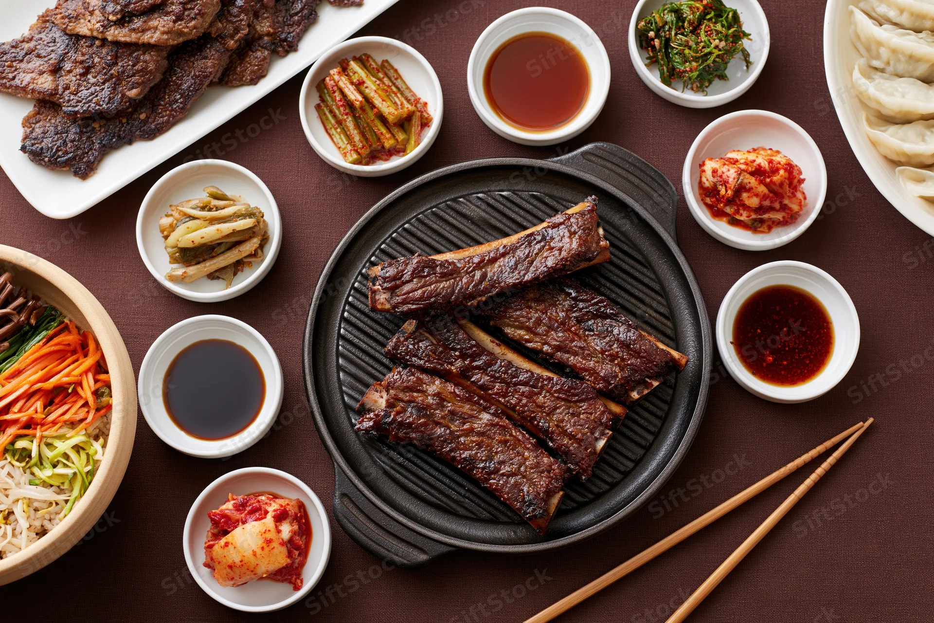 Overhead view of korean barbecue spread with grilled galbi ribs, side dishes, sauces, and chopsticks on a dark table