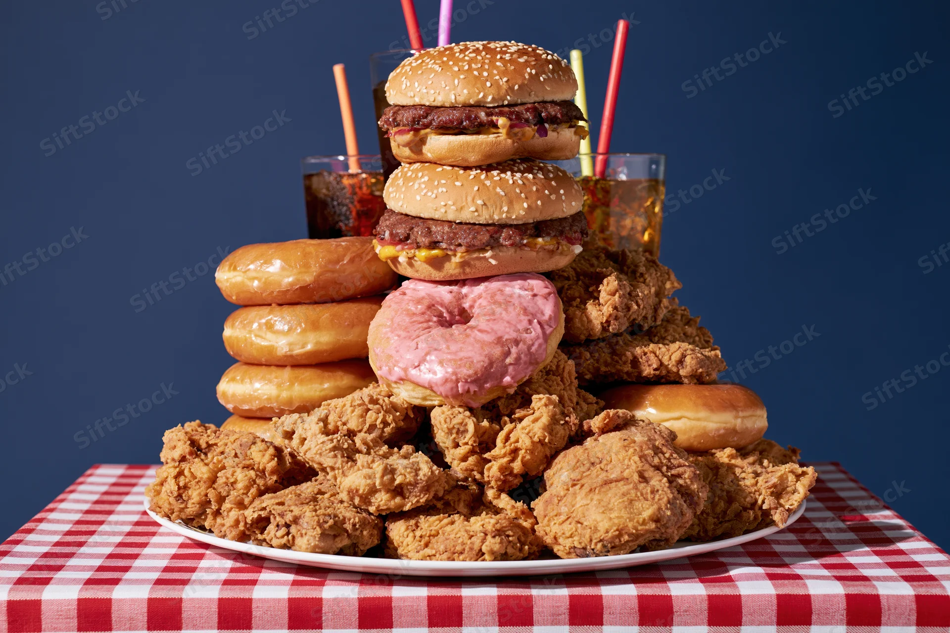 Pile of burgers, fried chicken, donuts and soda stacked on a plate on a red and white checkered tablecloth