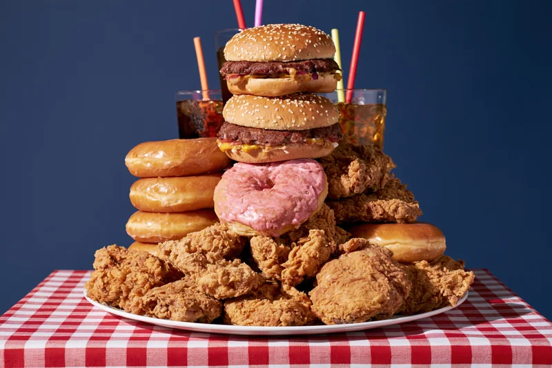 Pile of burgers, fried chicken, donuts and soda stacked on a plate on a red and white checkered tablecloth