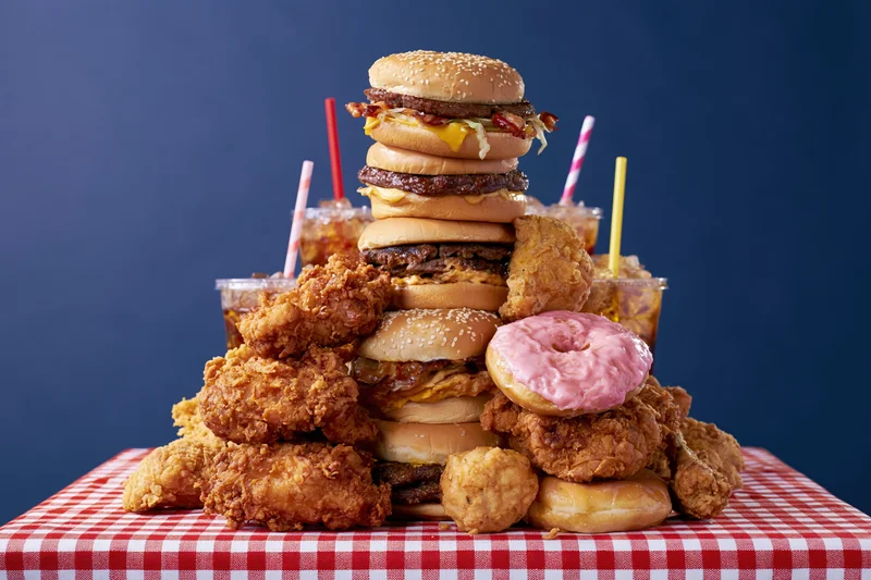 Towering pile of burgers, fried chicken, donuts and sodas on a red and white checkered tablecloth