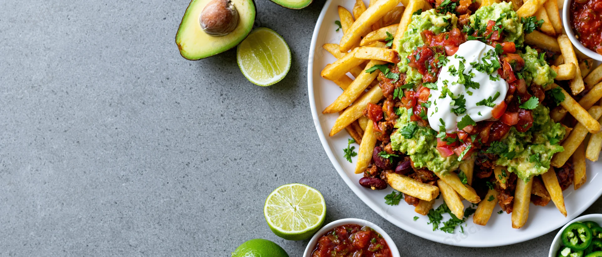 Loaded mexican-style fries topped with chili, guacamole, sour cream and salsa on a white plate with lime and avocado on gray surface