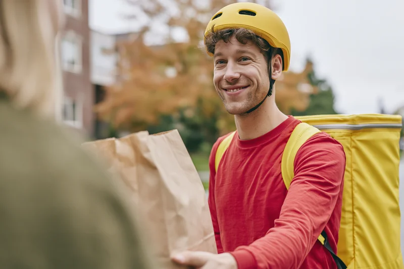 Smiling male courier handing paper bag to customer on city sidewalk