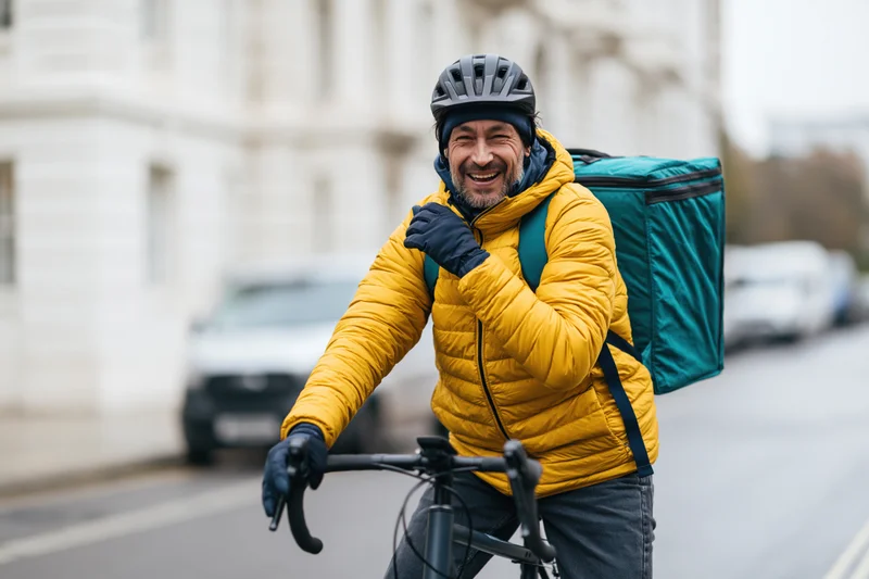 Smiling male delivery cyclist on bicycle in city street wearing yellow jacket and helmet with teal backpack