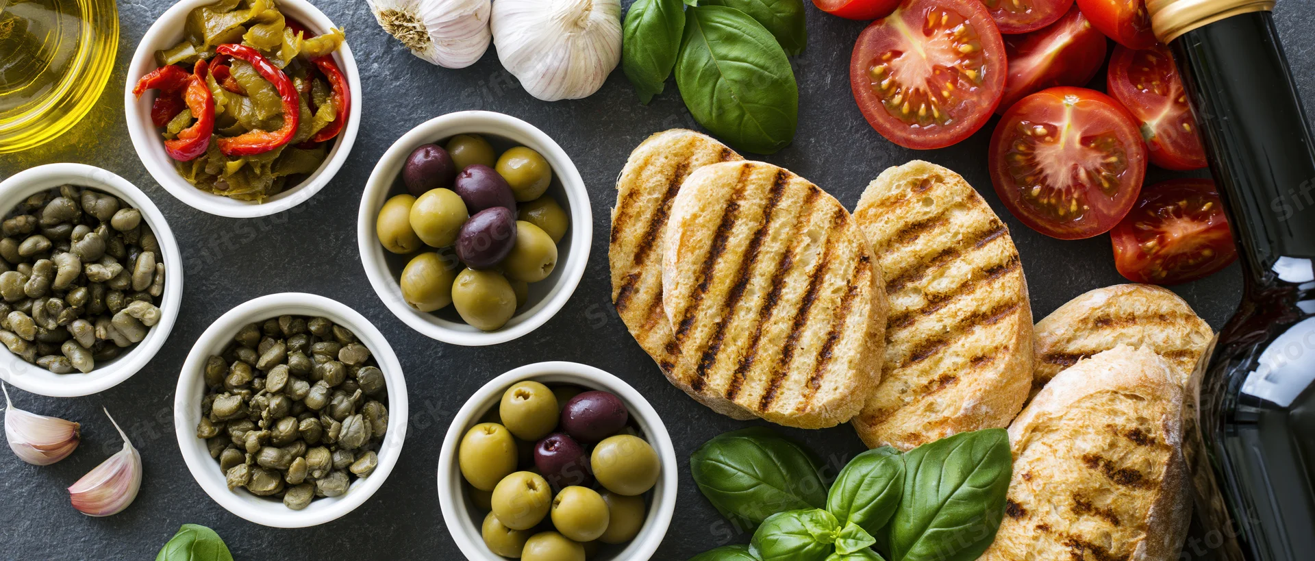 Mediterranean ingredients arranged on a dark slate surface with grilled bread, olives, tomatoes, basil, garlic, capers, and olive oil in small bowls