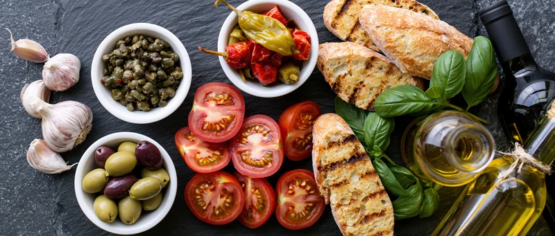 Mediterranean ingredients arranged on a dark slate surface including tomatoes, garlic, olives, capers, peppers, basil, bread, olive oil and wine
