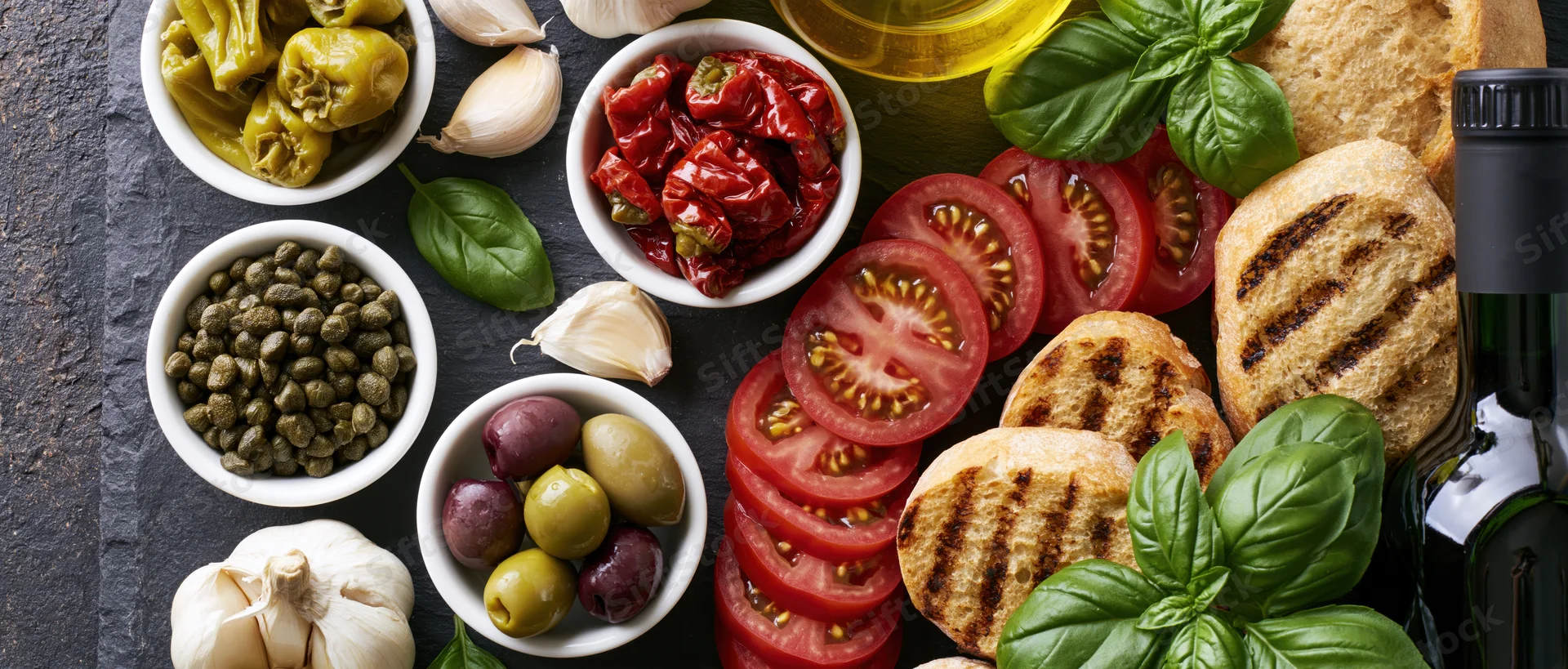 Mediterranean ingredients arranged on a dark slate surface including tomatoes, olives, garlic, basil, capers, bread, and olive oil