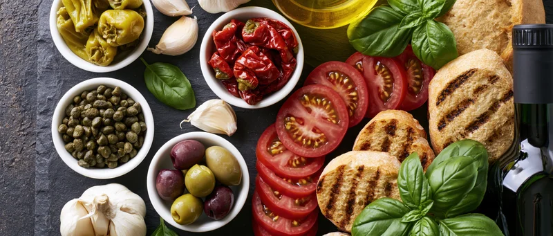 Mediterranean ingredients arranged on a dark slate surface including tomatoes, olives, garlic, basil, capers, bread, and olive oil