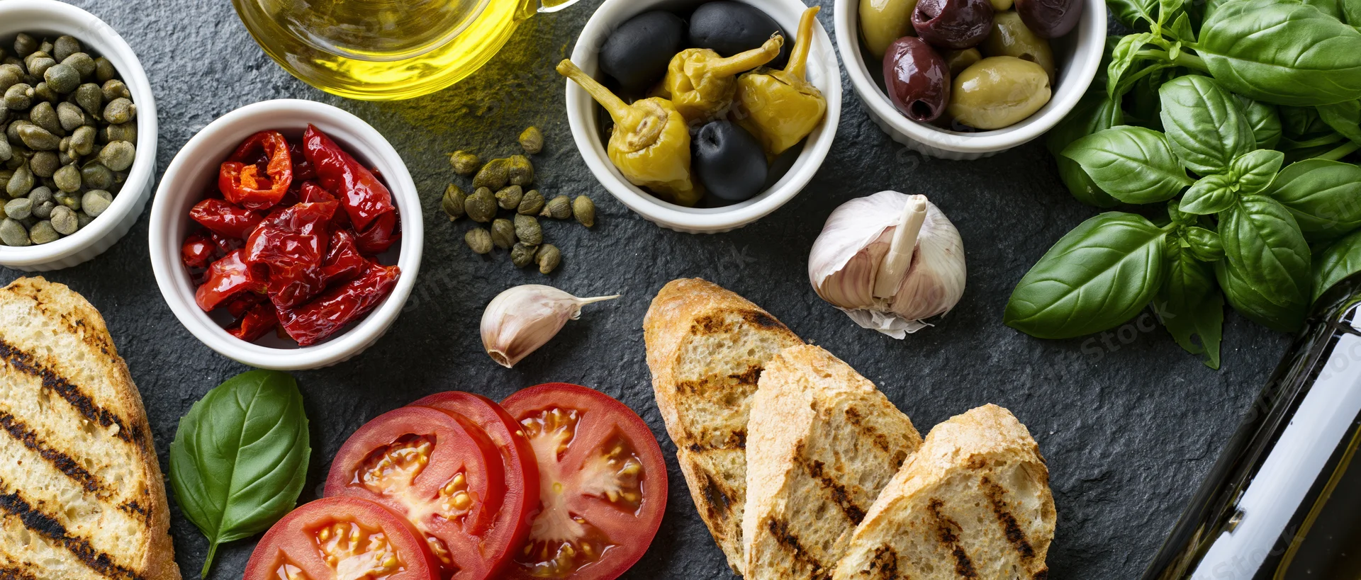 Mediterranean ingredients arranged on dark slate surface with grilled bread, tomatoes, basil, olives, capers and olive oil