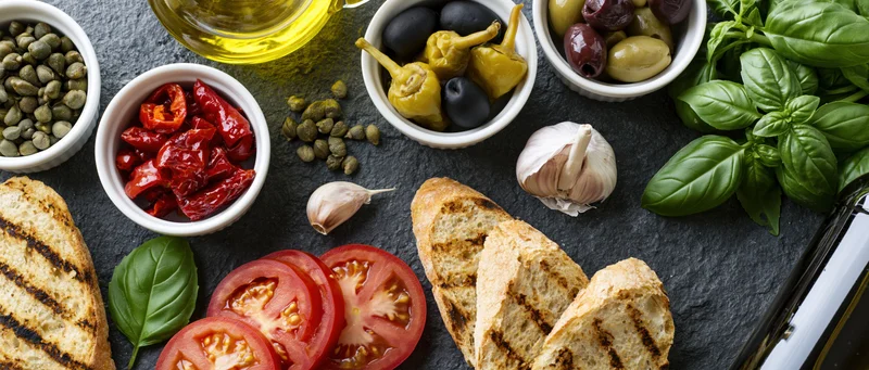 Mediterranean ingredients arranged on dark slate surface with grilled bread, tomatoes, basil, olives, capers and olive oil