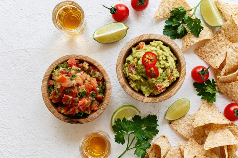 Mexican appetizer spread with guacamole and salsa in wooden bowls surrounded by tortilla chips lime and tequila on white textured surface