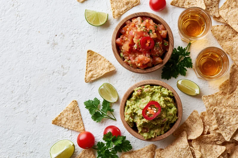 Mexican appetizer spread with guacamole and salsa in wooden bowls surrounded by tortilla chips and lime wedges on a white textured surface