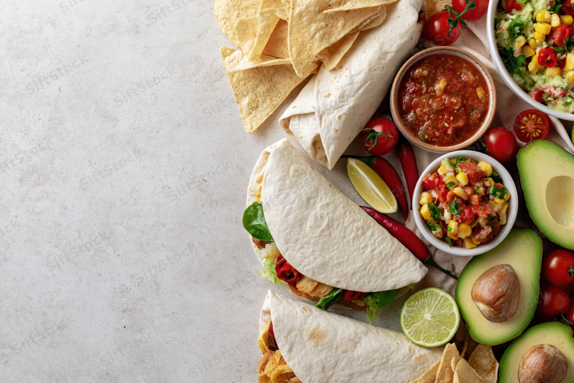 Mexican food spread with tacos, avocado, salsa and chips arranged along the right edge of a white surface
