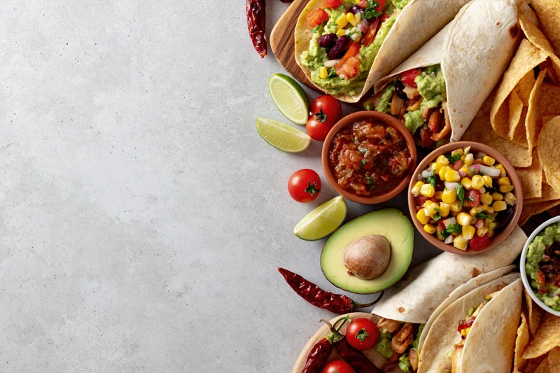 Mexican food spread with tacos, guacamole, salsa, and chips arranged along the right edge of a white surface