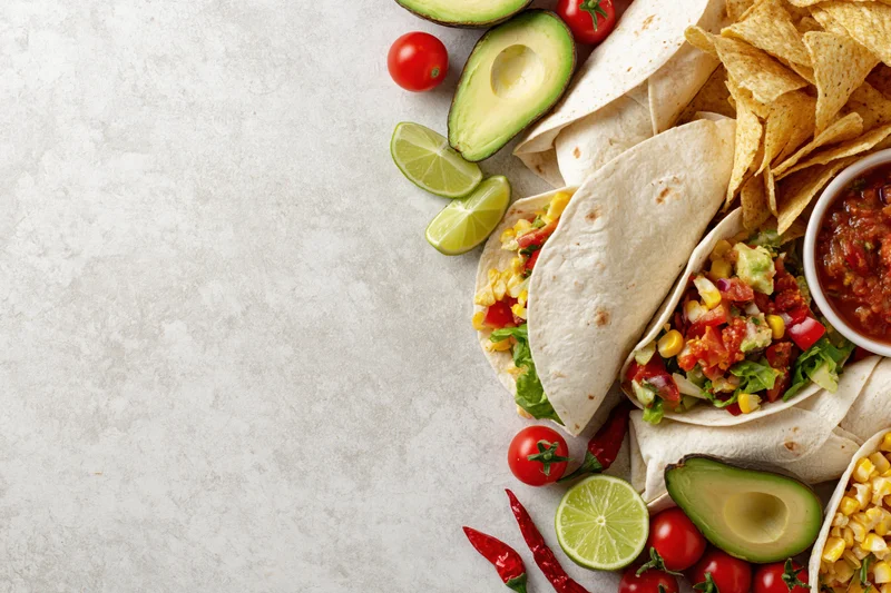 Mexican food spread with tacos, avocado, lime, tomato, and chips arranged along the right edge of a white surface