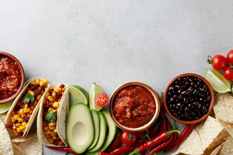 Mexican food spread with tacos, salsa, avocado, black beans and lime arranged along bottom edge of frame