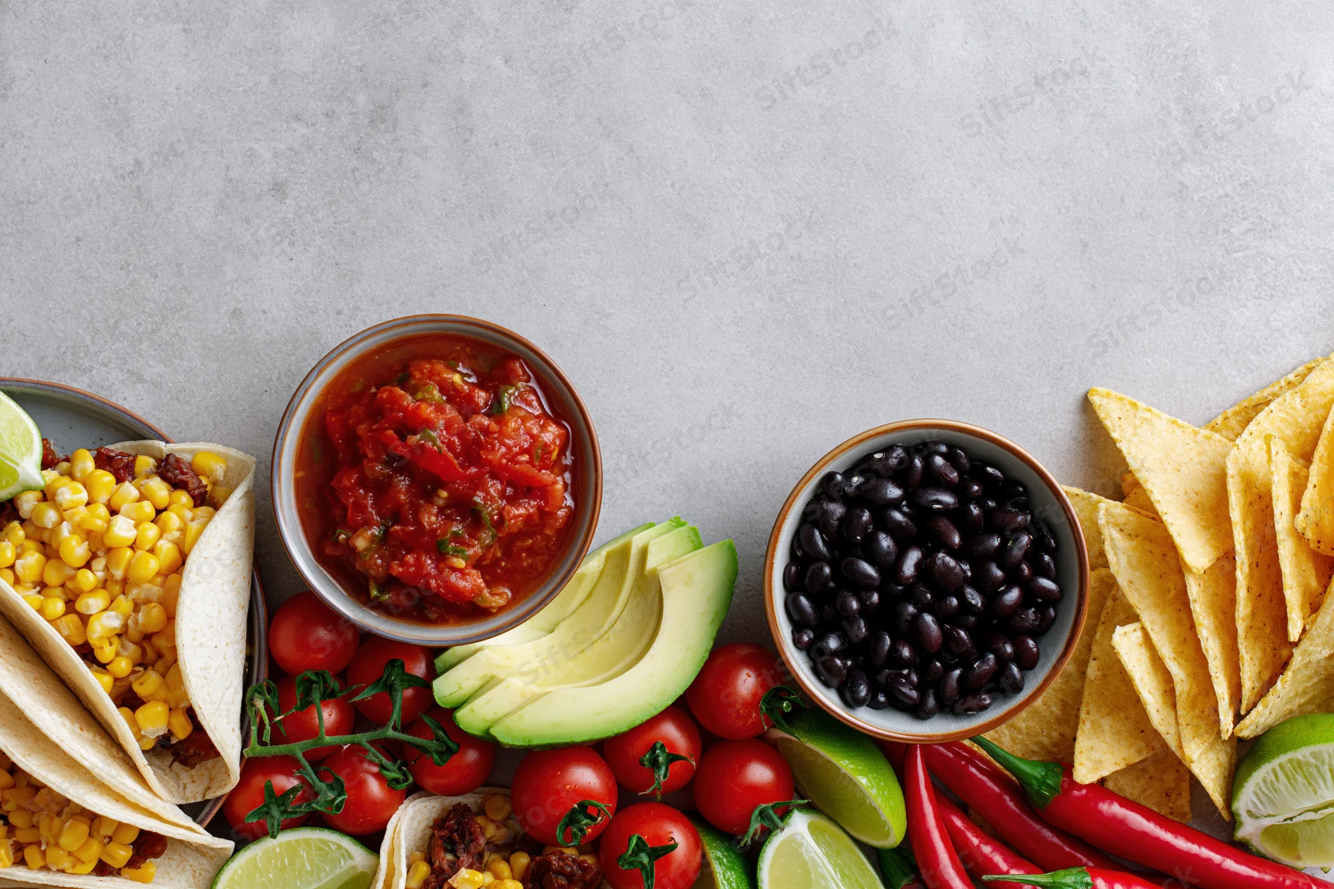 Mexican food spread with tacos, salsa, avocado, black beans, tomatoes, lime, chili, and chips arranged along bottom edge of gray surface