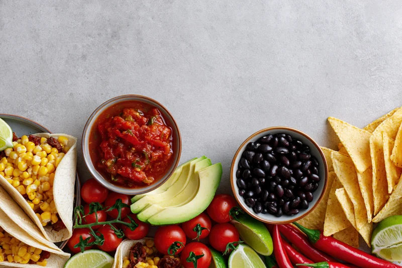 Mexican food spread with tacos, salsa, avocado, black beans, tomatoes, lime, chili, and chips arranged along bottom edge of gray surface