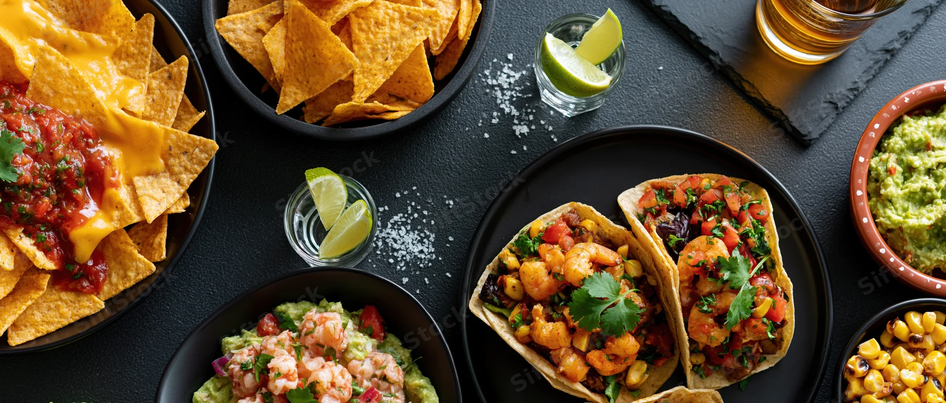 Mexican food spread with tacos, nachos, guacamole and beer arranged on a dark stone table in overhead view