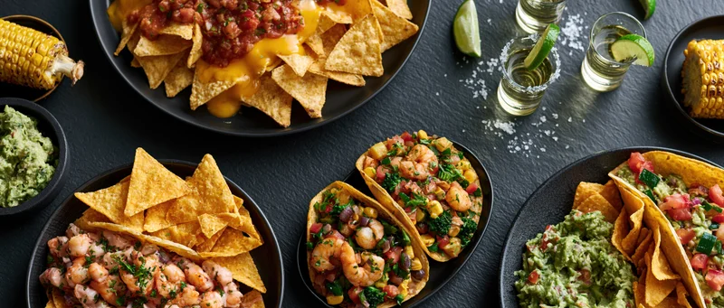 Mexican food spread with tacos, nachos, guacamole, shrimp, corn, and tequila shots on a dark stone table
