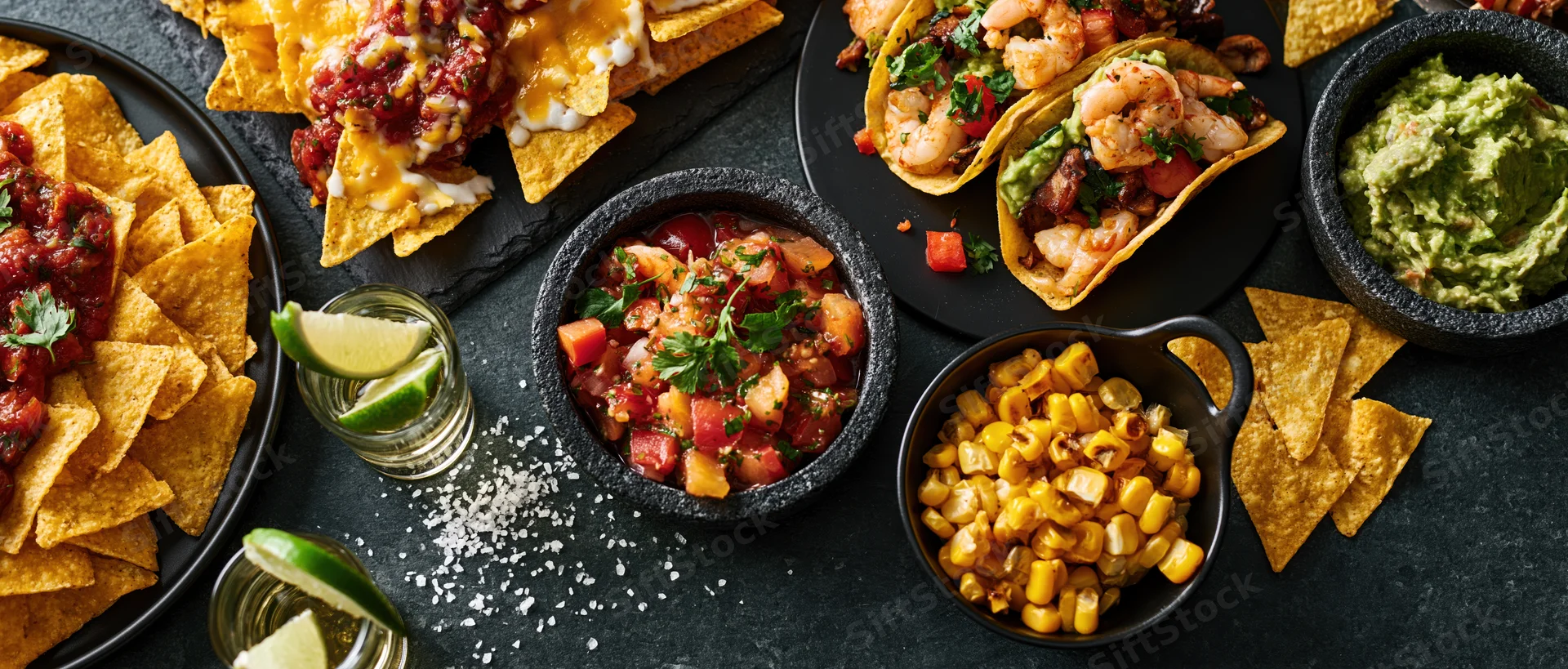 Mexican food spread with tacos, nachos, guacamole, salsa, and tequila on dark stone surface