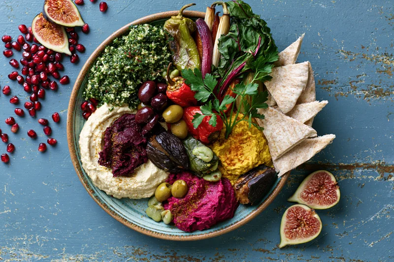 Overhead view of a middle eastern mezze platter with hummus, beet dip, figs, pomegranate, olives, peppers, parsley, and flatbread on a blue rustic surface