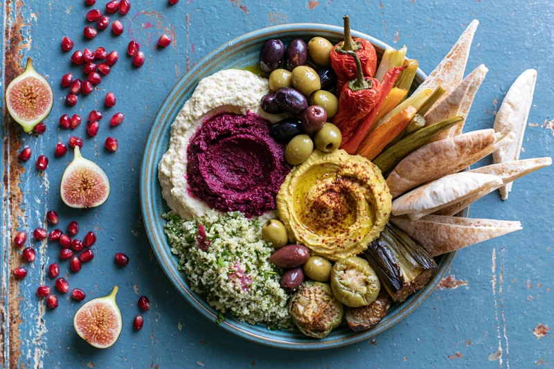 Overhead view of a middle eastern mezze platter with hummus, olives, pita, vegetables, figs and pomegranate seeds on a blue rustic surface