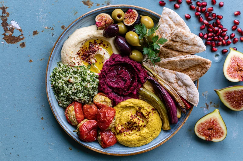 Overhead view of a middle eastern mezze platter with hummus, pita, olives, figs and pomegranate seeds on a blue textured surface