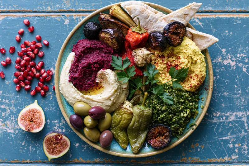 Overhead view of a middle eastern mezze platter with hummus, beet dip, tabbouleh, pita, figs, olives, and pomegranate seeds on a blue wooden table