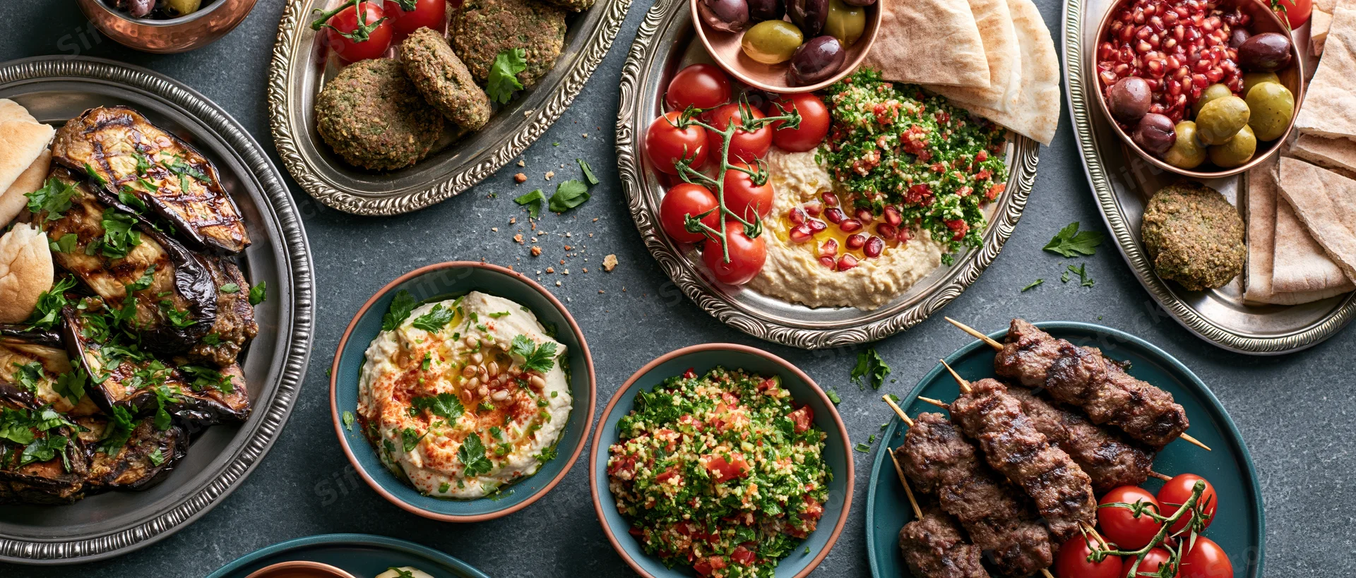 Middle eastern mezze spread with hummus, falafel, kebabs and vegetables arranged on a dark slate table in overhead view
