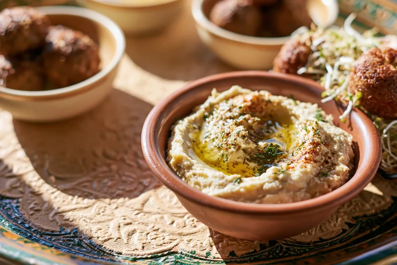 Hummus in a clay bowl garnished with olive oil and herbs on a decorated metal tray with mezze dishes around