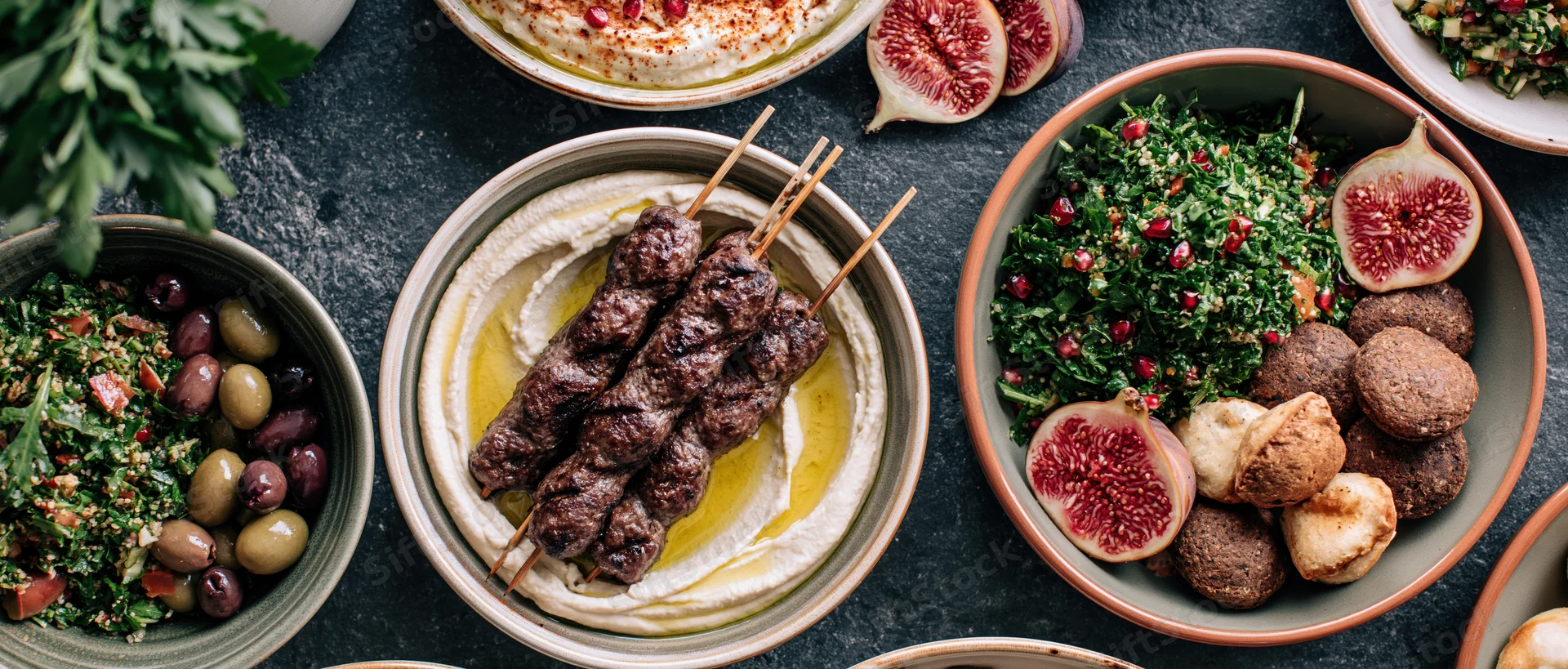 Overhead view of a middle eastern mezze spread with hummus, kebabs, figs, olives and salads on a dark table