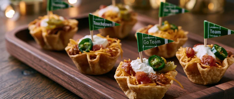 Mini taco cups filled with cheese, tomatoes, jalapenos, and sour cream on a wooden tray with football flags for a game-day party