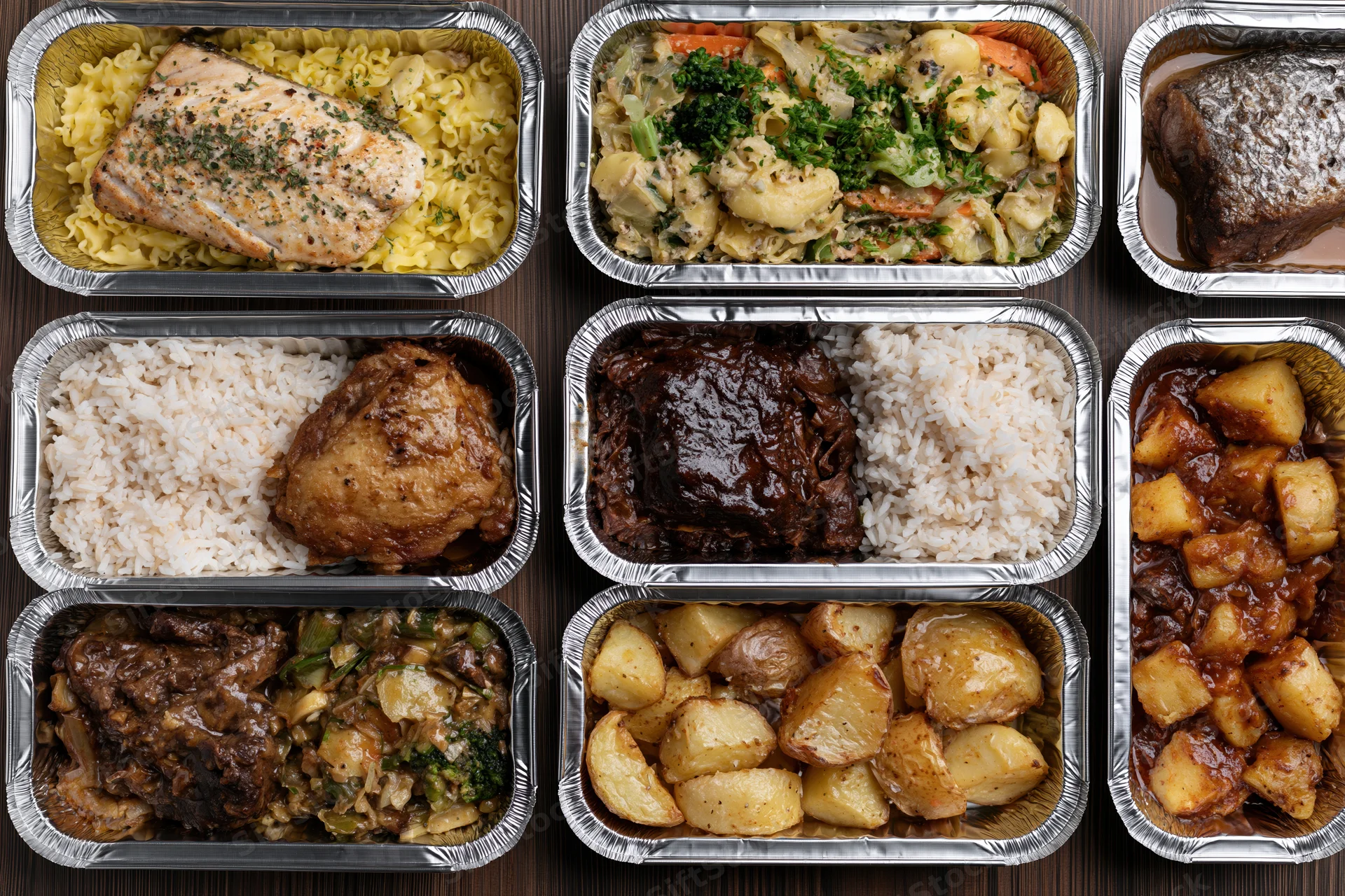 Multiple aluminum foil takeout containers arranged in grid on dark wood surface, each filled with different cooked meals including fish, chicken, beef, potatoes, rice, and pasta